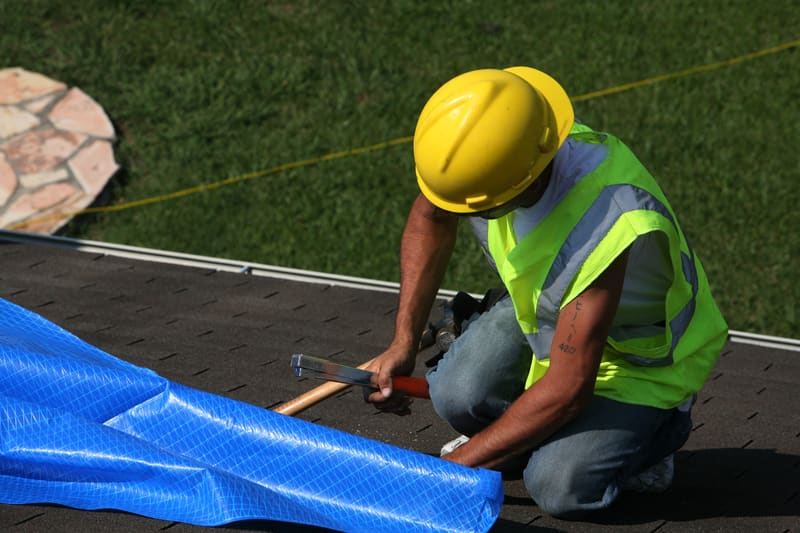 A worker in a hard hat and high-visibility vest fastening a protective cover over a roof